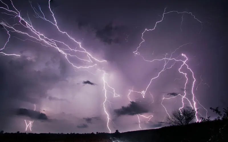 Dramatic violet and white lightning branching across a dark stormy sky above a silhouette of trees and hills