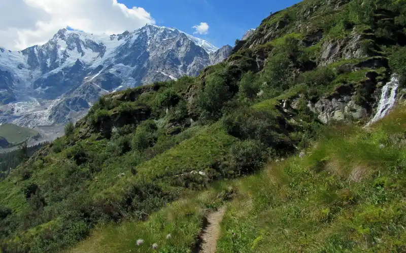 Narrow dirt trail climbing a green mountain slope beside a small waterfall, with a glacier-covered massif in the background
